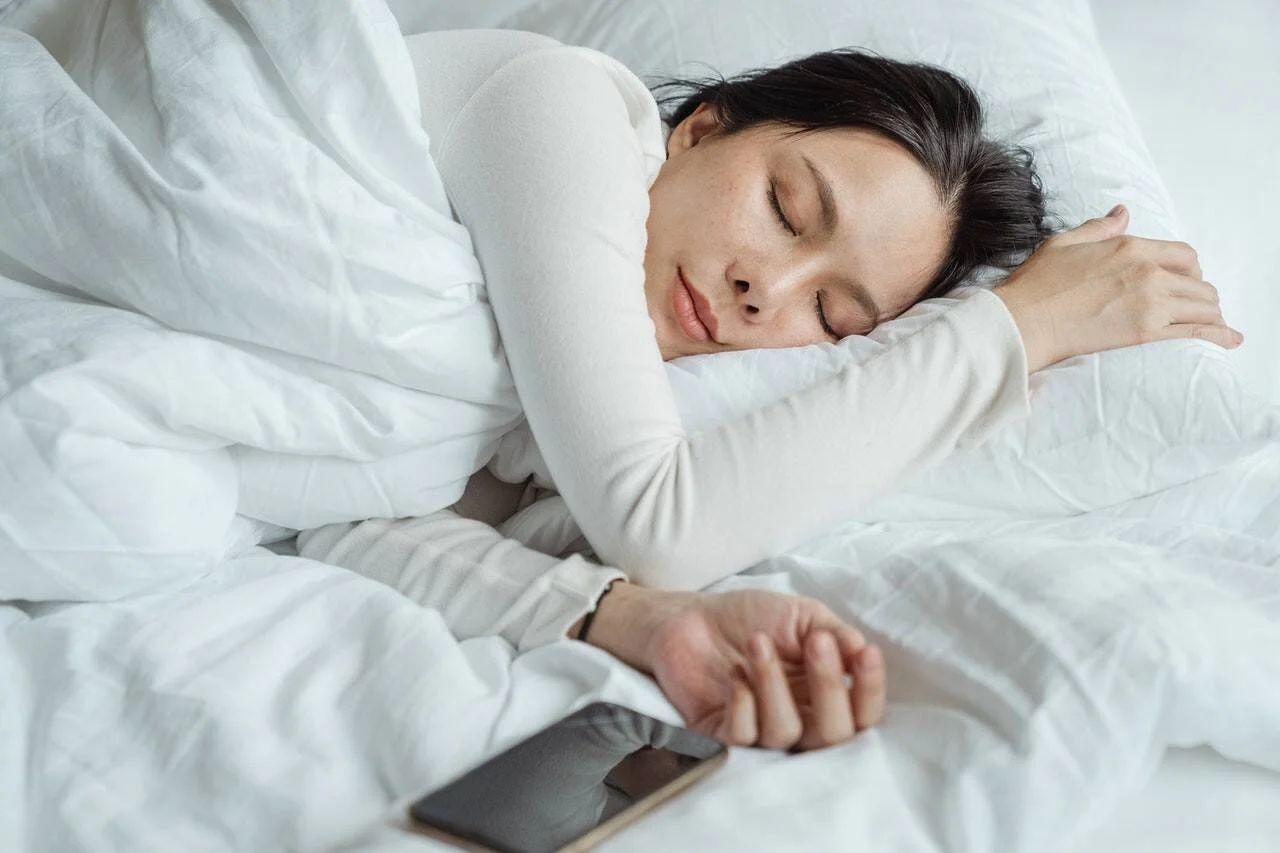Mujer joven dormida en una cama con ropa blanca, con un smartphone apoyado junto a su mano.