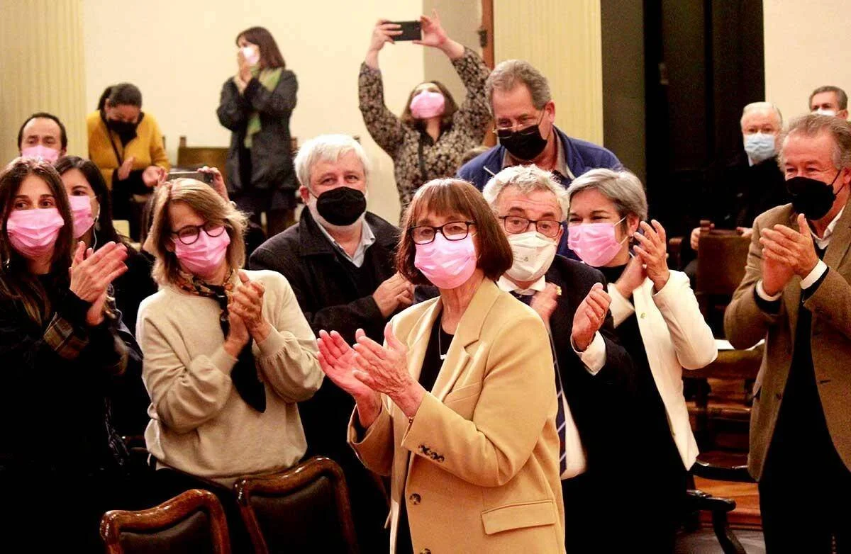 Grupo de personas con mascarillas rosas y negras aplauden de pie en una sala de actos durante un evento.