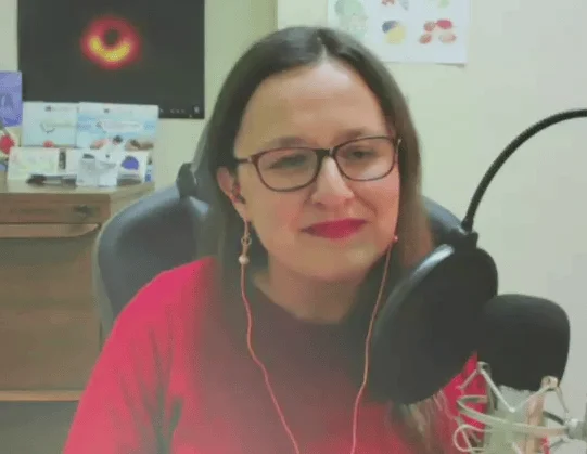 Mujer con anteojos y remera roja frente a un micrófono en un espacio de grabación doméstico con póster de agujero negro al fondo.