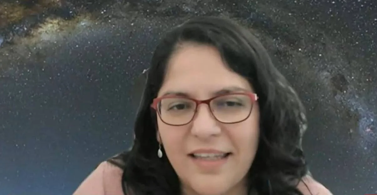 Mujer joven con anteojos rojos sonríe frente a un fondo virtual de la Vía Láctea en videollamada.