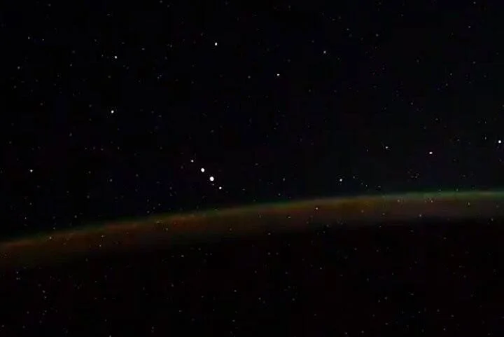 Franja luminosa verde y naranja de una aurora boreal sobre la curva de la Tierra, fotografiada desde el espacio con estrellas al fondo.