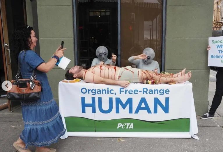 Activistas de PETA disfrazados de alienígenas simulan comer a un humano en una mesa con el cartel 'Organic, Free-Range Human' en la calle.