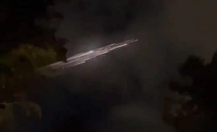 Objeto luminoso con estela brillante atraviesa el cielo oscuro nocturno entre siluetas de árboles.