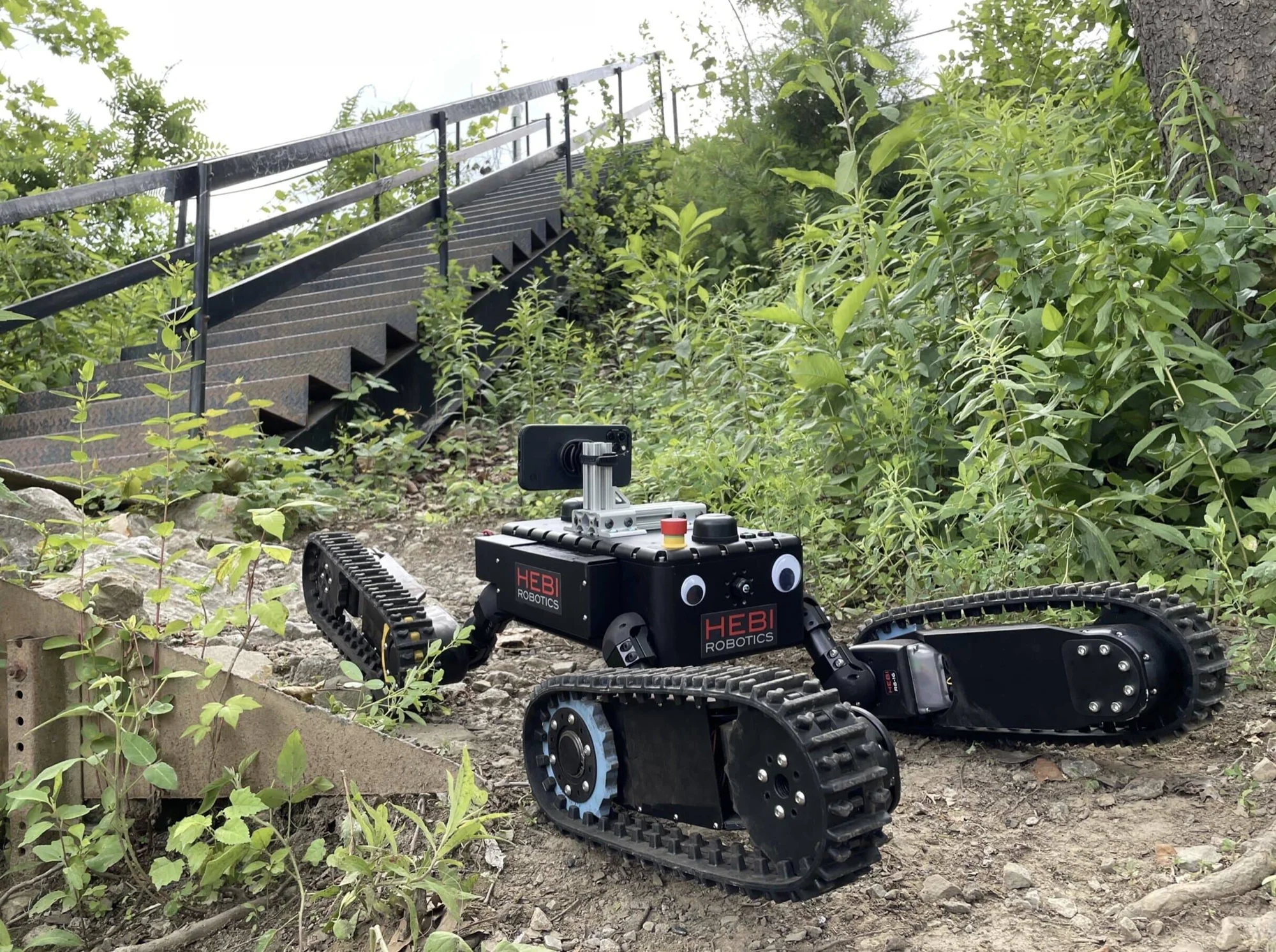 Robot de orugas HEBI Robotics navegando por terreno irregular con vegetación y escaleras metálicas al fondo.