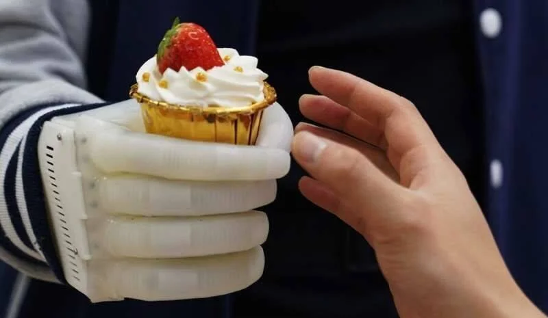 Una mano protésica robótica blanca sostiene un cupcake con crema y fresa mientras una mano humana se acerca a tomarlo.