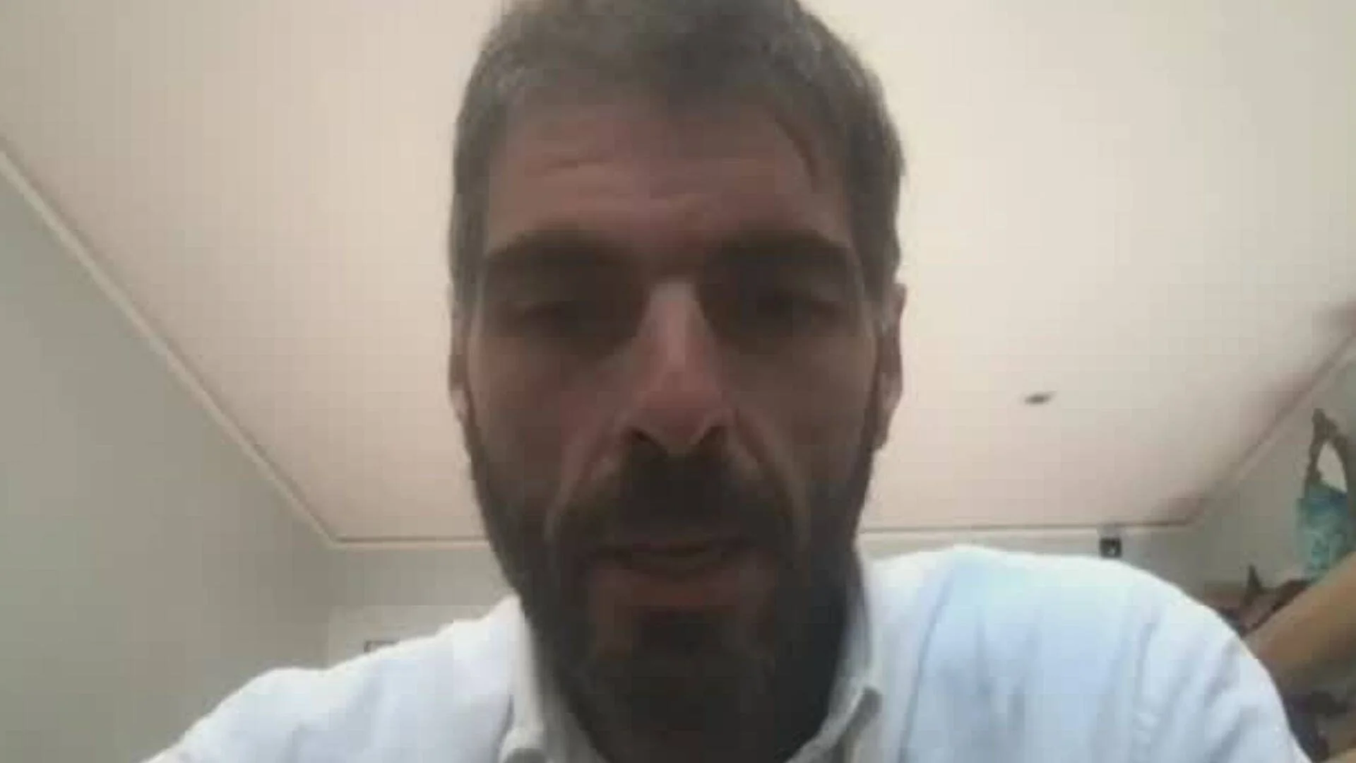 Hombre adulto con barba oscura y camisa blanca habla frente a una cámara en un interior.