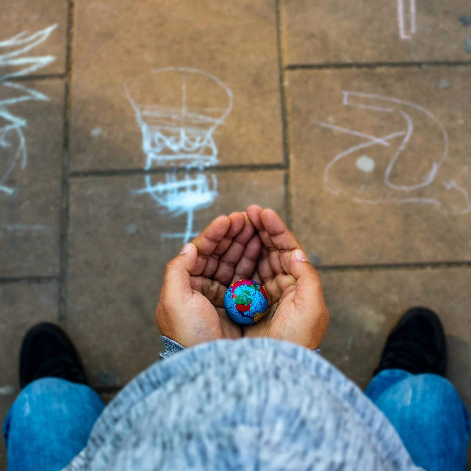 Persona sostiene con ambas manos un pequeño globo terráqueo de colores sobre un suelo de adoquines con dibujos de tiza.