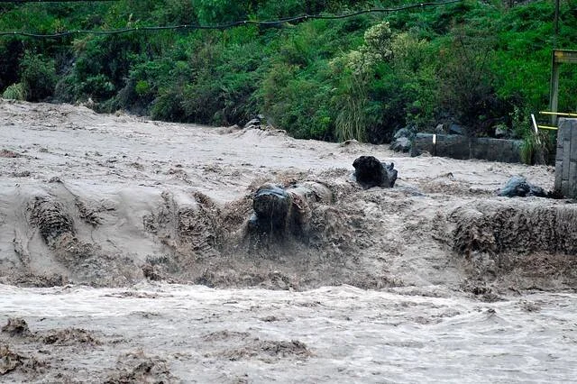 Río desbordado con corriente turbia y lodosa arrastrando rocas, con vegetación verde al fondo.