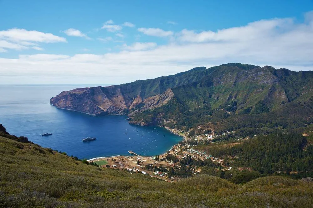 Vista aérea de una bahía de aguas turquesas rodeada de montañas verdes con un pequeño poblado costero y embarcaciones ancladas.