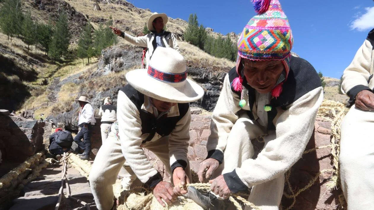 Hombres con vestimenta andina tradicional trenzando cuerdas de paja en un paisaje de montaña andina.