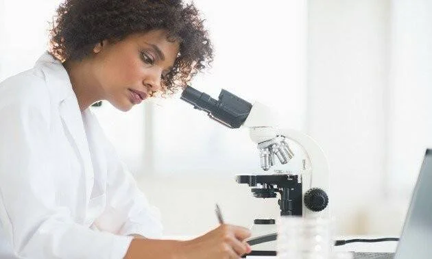 Mujer con bata blanca observa por un microscopio mientras toma notas en un laboratorio.