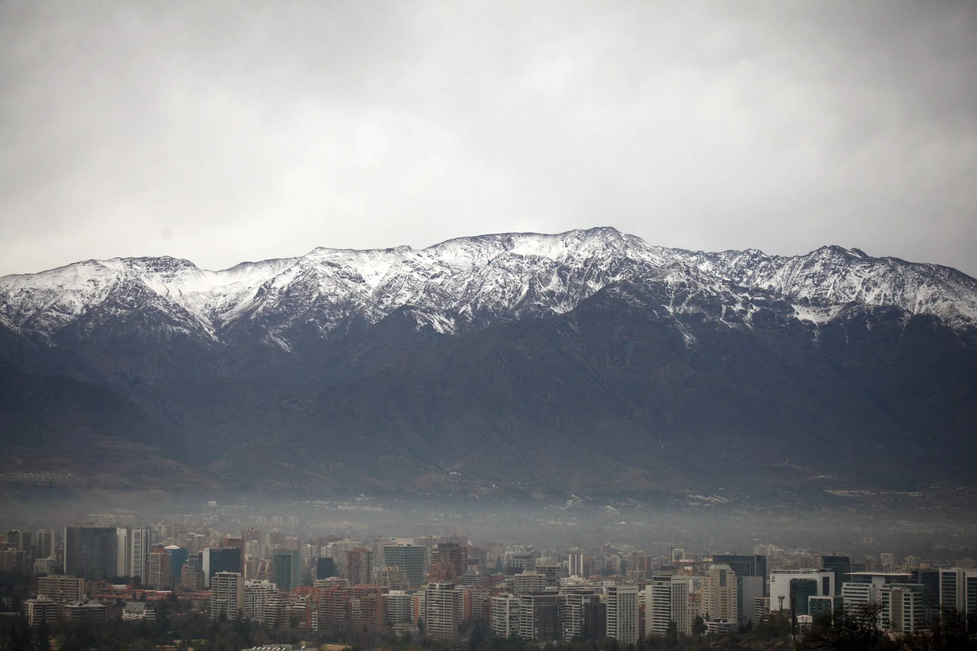 Skyline de una ciudad con edificios envueltos en smog y una cordillera con cumbres nevadas bajo cielo nublado.