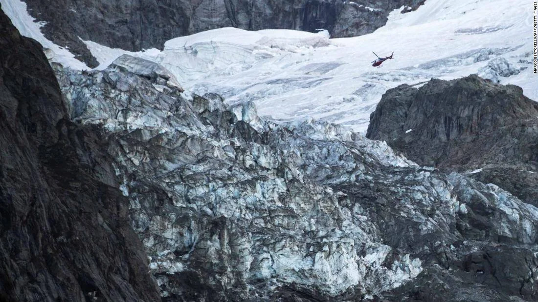 Un helicóptero sobrevuela la zona rocosa y glacial de una alta montaña cubierta de nieve y hielo.
