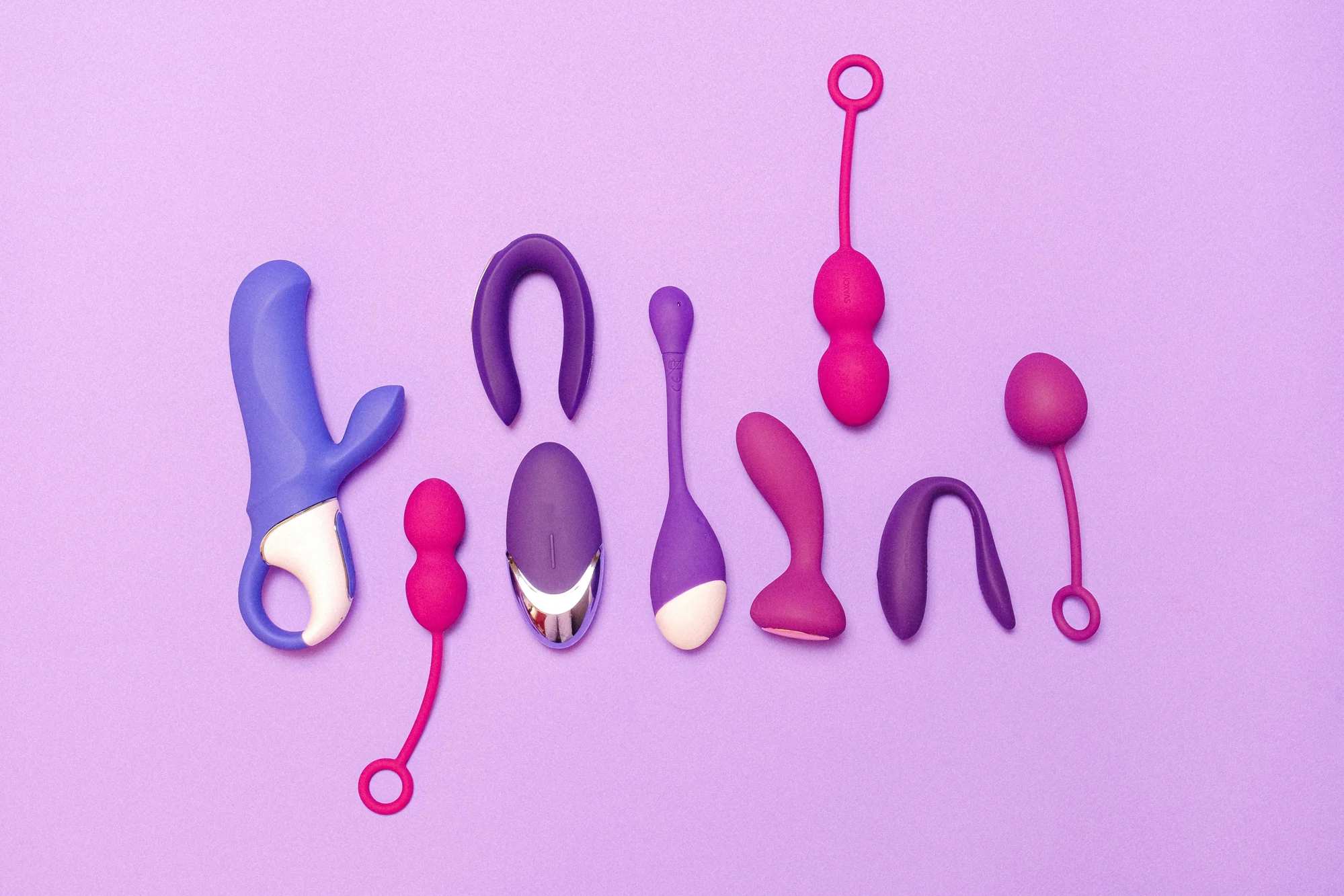 Colección de juguetes sexuales de silicona en tonos azul, morado y rosa dispuestos sobre fondo lila.