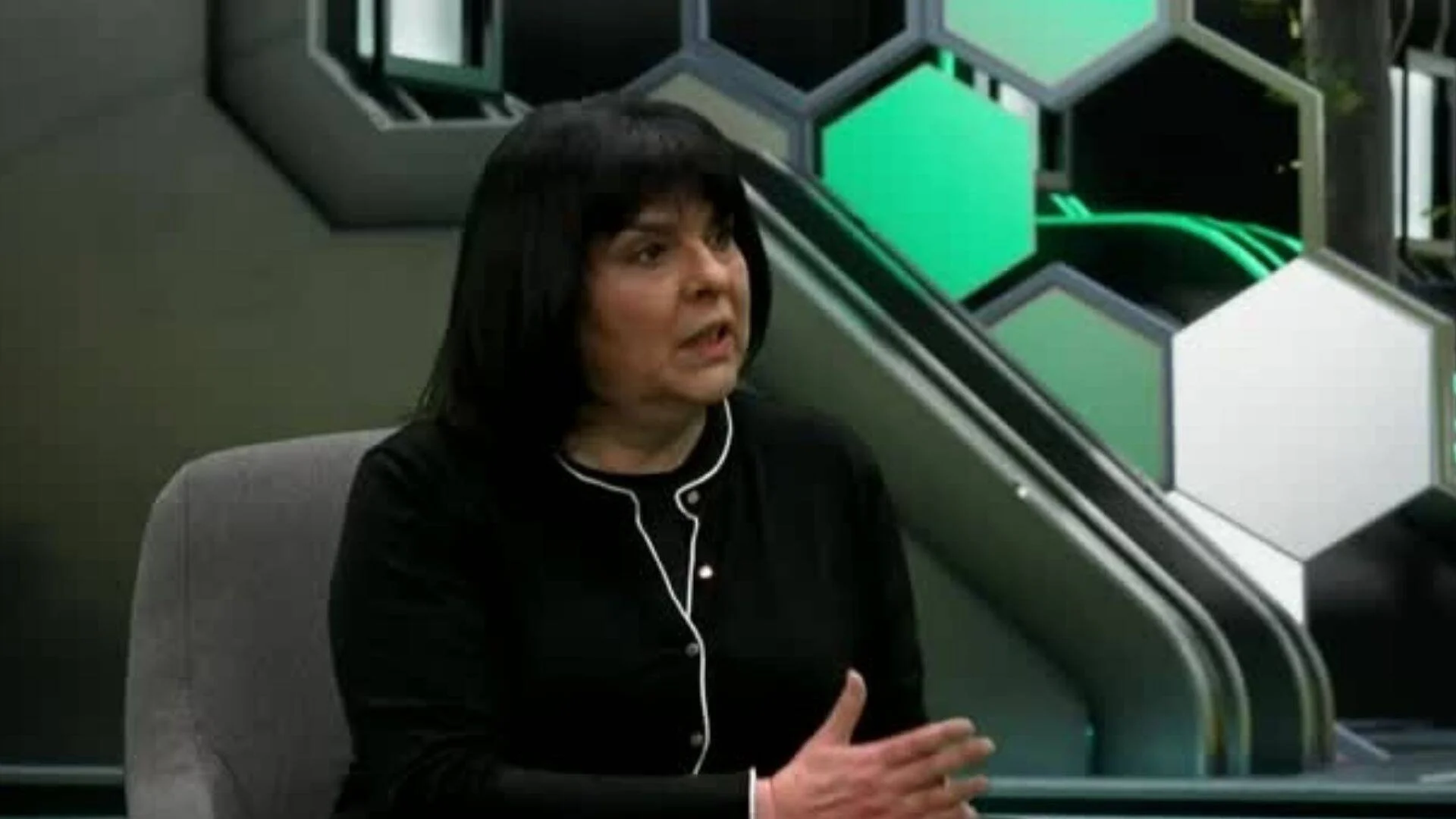 Mujer de cabello negro hablando en un estudio de televisión con decorado de paneles hexagonales verdes y grises.
