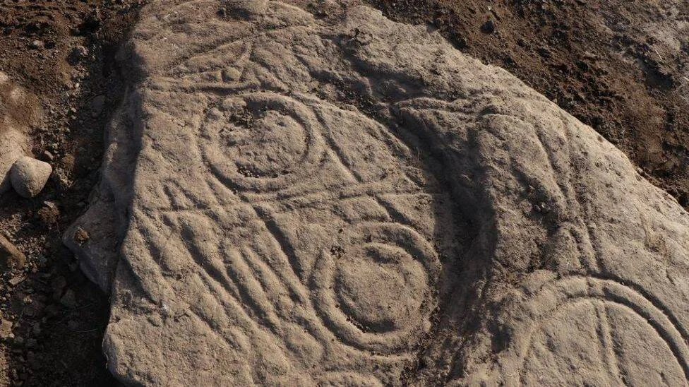 Roca plana con grabados prehistóricos en espiral y formas curvilíneas, parcialmente enterrada en tierra.