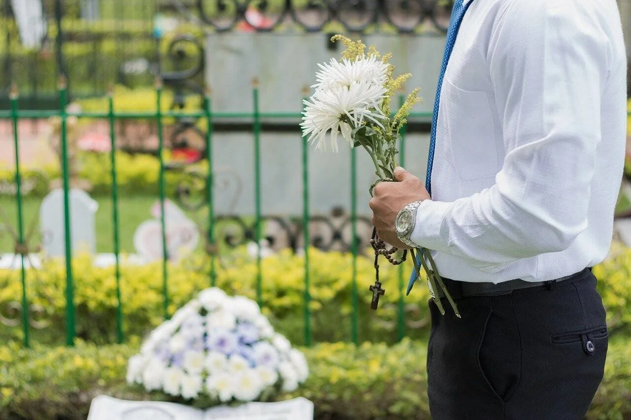 Hombre vestido de luto sostiene flores blancas y un rosario frente a una tumba en un cementerio.