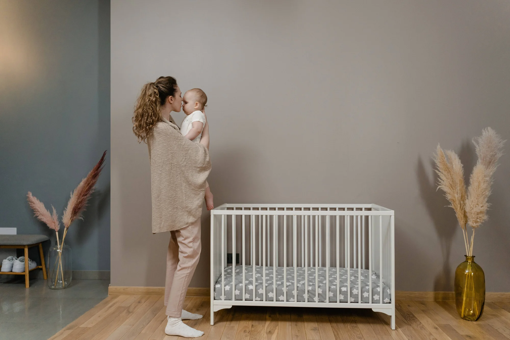 Madre besa a su bebé en brazos junto a una cuna blanca en una habitación infantil decorada con plantas de pampa.