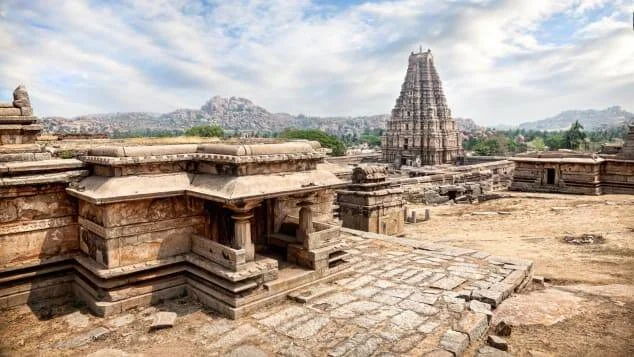 Complejo de ruinas de piedra con torre gopuram de templo hindú en Hampi, rodeado de paisaje rocoso bajo cielo nublado.
