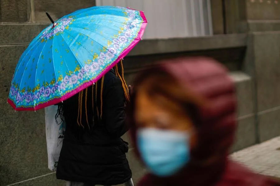 Una persona con paraguas azul floreado y otra con mascarilla y capucha roja caminan en un día lluvioso.