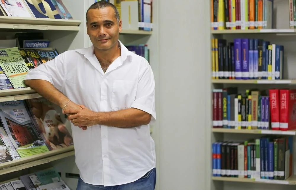 Hombre con camisa blanca apoyado en un expositor de revistas científicas en una biblioteca con estantes de libros al fondo.