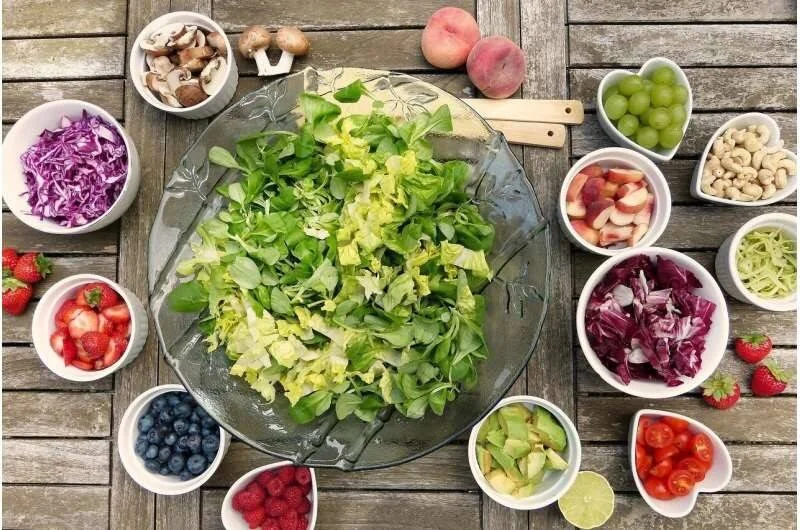 Cuenco grande de ensalada verde rodeado de bowls con frutas, verduras y frutos secos sobre mesa de madera.