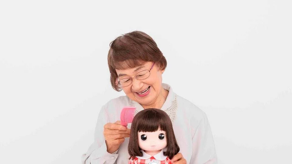 Anciana sonriente cepilla el cabello de una muñeca robótica de niña con ojos grandes, sobre fondo blanco.