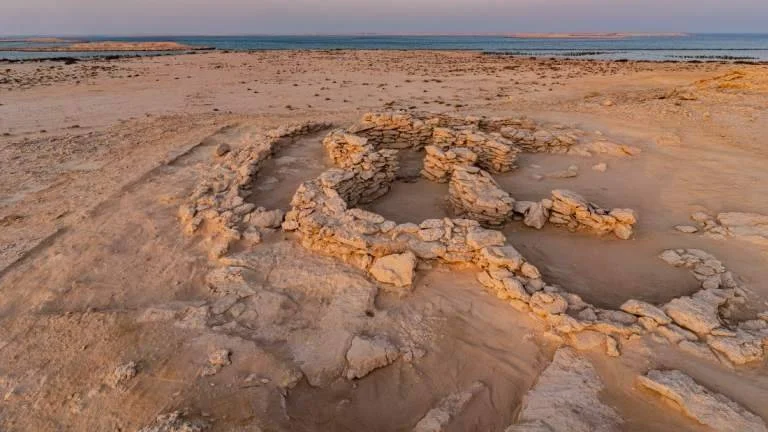 Restos de muros de piedra de una estructura antigua en un paisaje desértico junto al mar, fotografiados al atardecer.