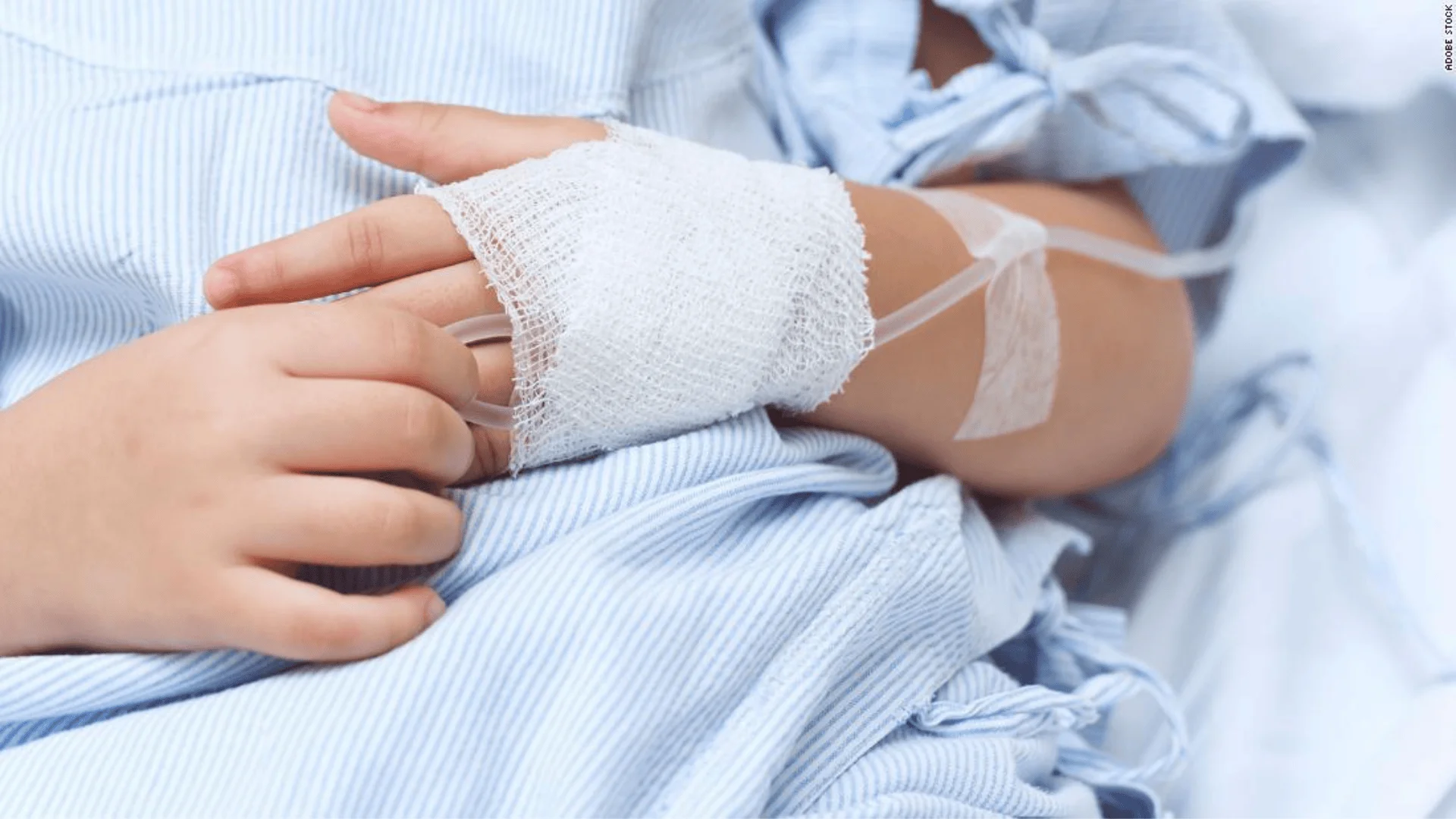 Mano de un niño con venda y catéter intravenoso colocado, sobre ropa de cama azul de hospital.