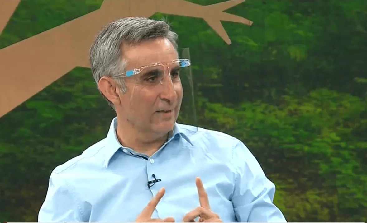 Hombre de cabello canoso con lentes y camisa celeste habla gesticulando en un estudio de televisión con fondo verde.