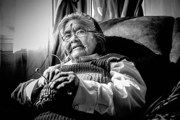 Mujer mayor con anteojos teje tranquilamente sentada en un sillón, en fotografía en blanco y negro.