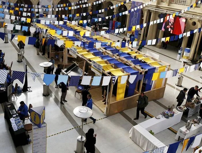 Vista aérea de una feria o exposición en un salón interior decorado con banderines azules y amarillos, con stands de madera y visitantes recorriendo el lugar.