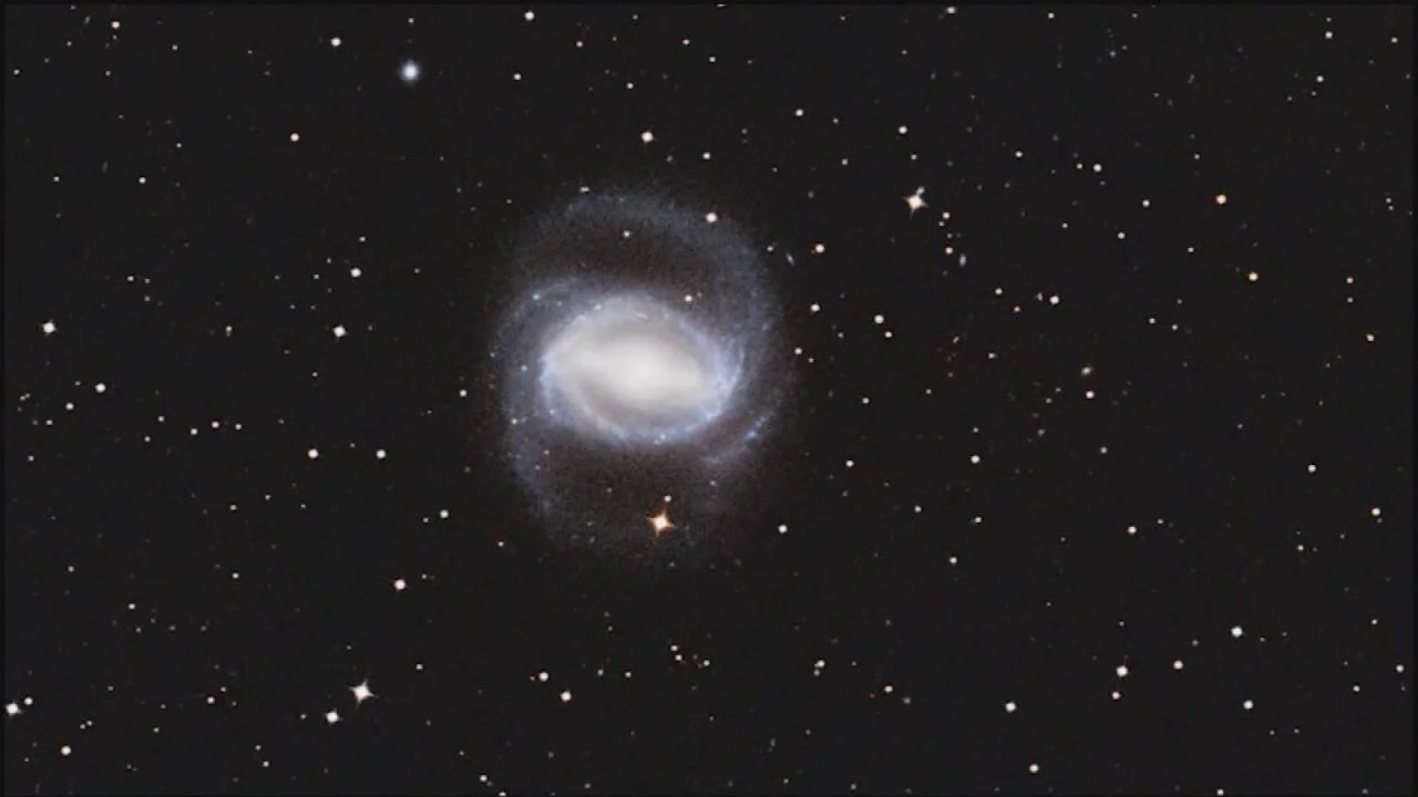 Galaxia espiral barrada con núcleo luminoso blanco y brazos azulados rodeada de estrellas sobre fondo negro del espacio.