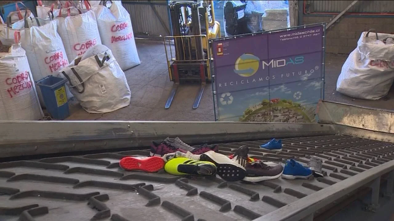 Zapatillas deportivas usadas sobre una cinta transportadora en planta de reciclaje Midas, con sacos de material y un montacargas al fondo.