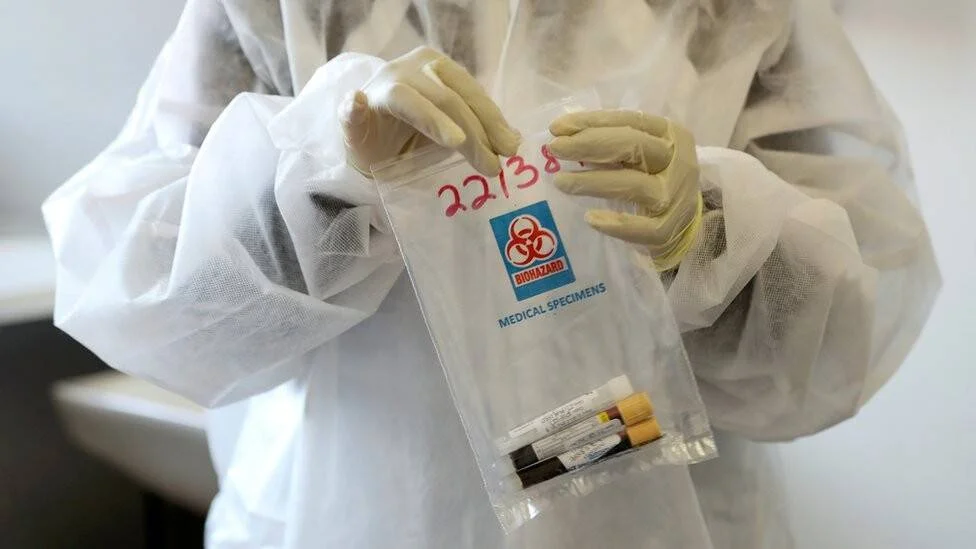Profesional de salud con traje y guantes sostiene una bolsa de riesgo biológico con muestras médicas en su interior.