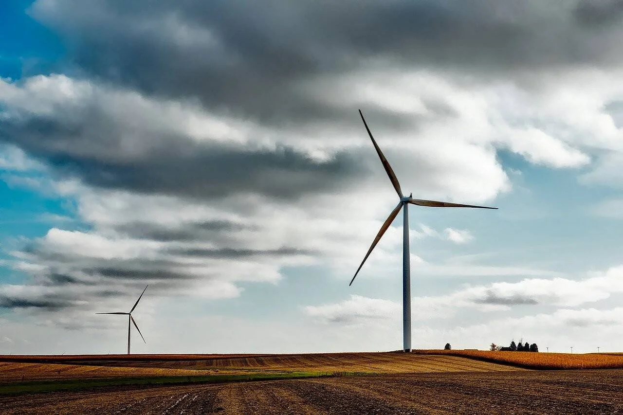 Dos aerogeneradores se elevan sobre campos de cultivo bajo un cielo parcialmente nublado, generando energía eólica.
