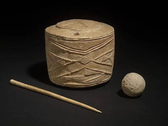 Cilindro de piedra neolítico con grabados geométricos, junto a una aguja de hueso y una pequeña esfera de piedra.