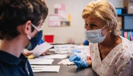 Mujer mayor con mascarilla y guantes azules conversa con un joven en una mesa de trabajo durante la pandemia.