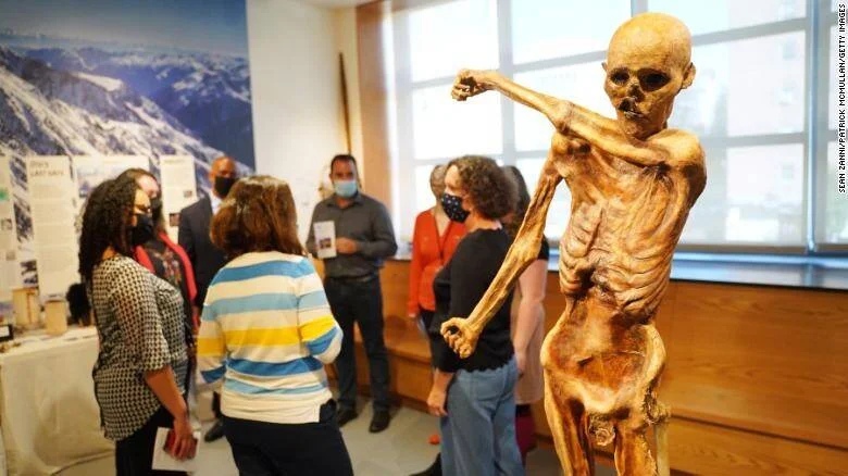 Visitantes con mascarillas observan una réplica de momia prehistórica expuesta en una sala de museo.