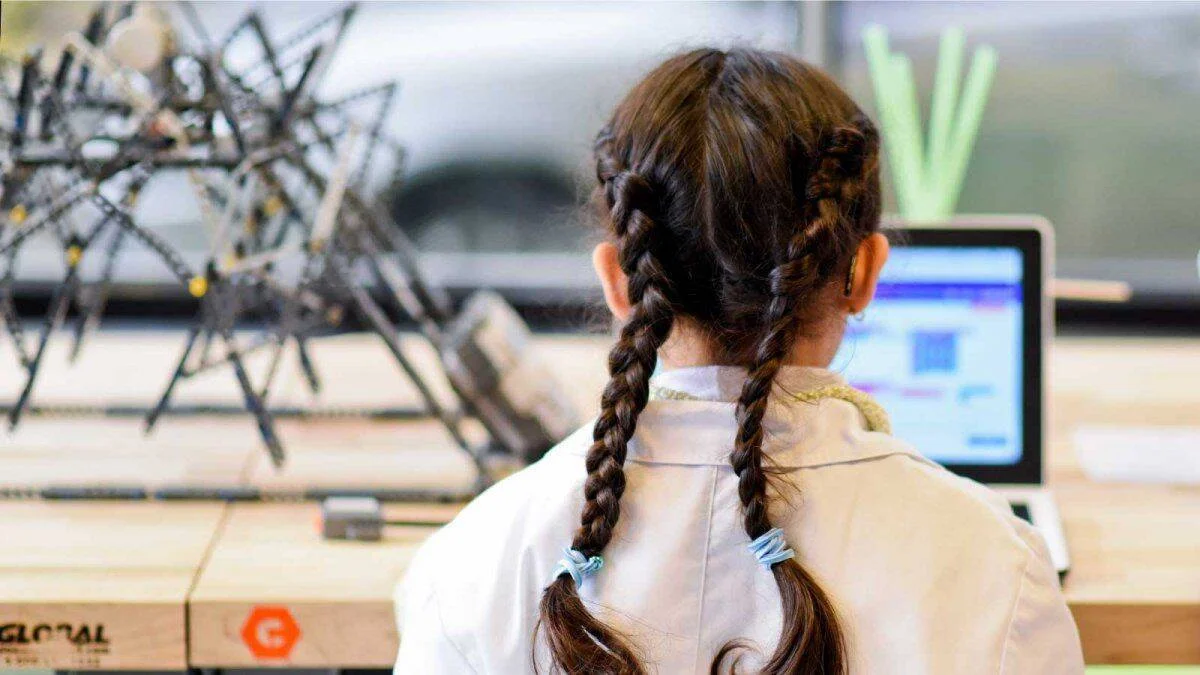 Niña vista de espaldas frente a una laptop y una estructura metálica de robótica en un taller educativo.