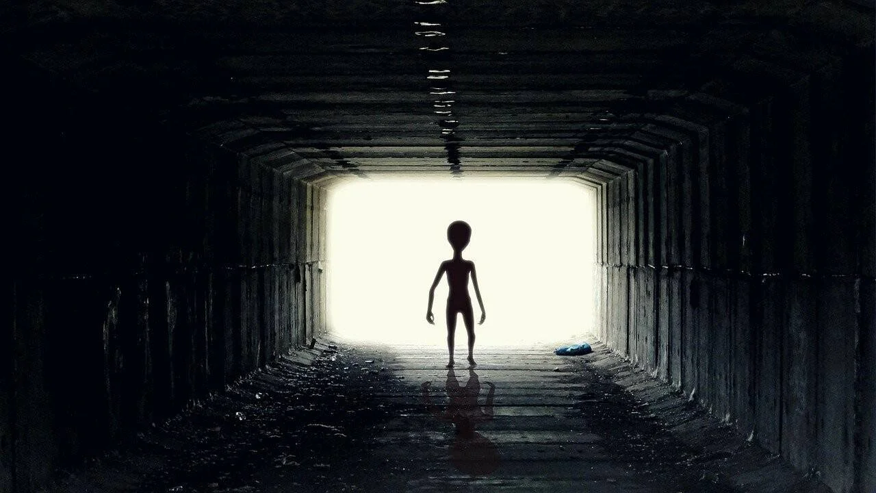 Silueta de figura extraterrestre de cabeza grande parada en un túnel oscuro con luz brillante al fondo.