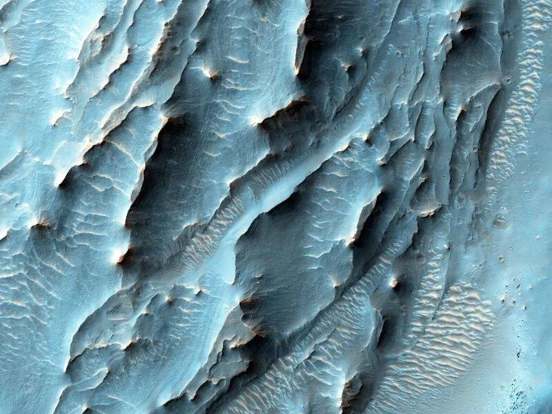 Vista aérea de la superficie marciana con formaciones rocosas estratificadas en tonos azul claro y gris oscuro.