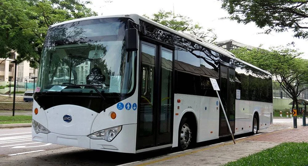 Autobús eléctrico de la marca BYD, blanco con techo negro, estacionado en una calle arbolada de zona urbana.
