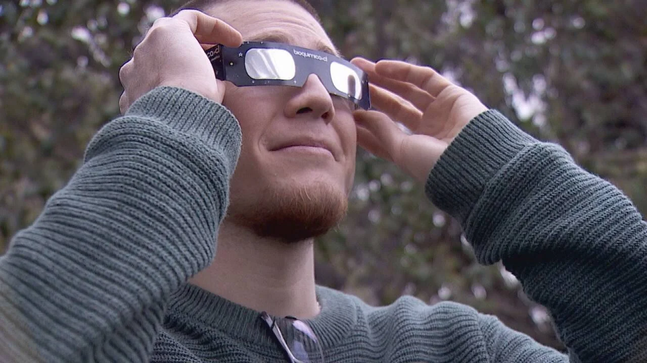 Hombre joven con lentes de cartón para eclipse solar mira hacia el cielo al aire libre.