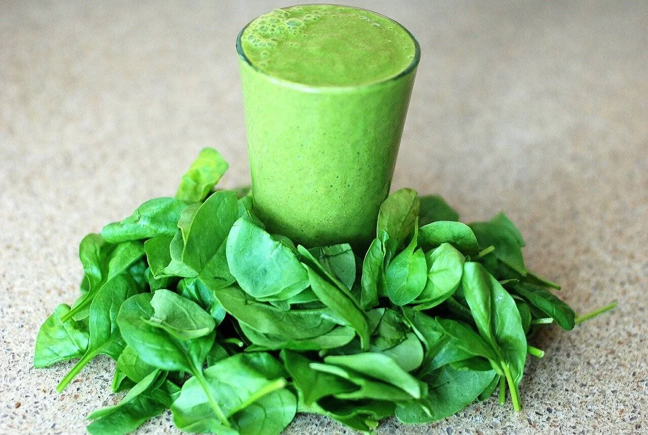 Vaso con batido verde rodeado de hojas frescas de espinaca sobre una superficie de piedra.