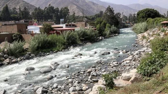 Río de aguas turbulentas flanqueado por rocas, vegetación y construcciones urbanas con montañas al fondo.