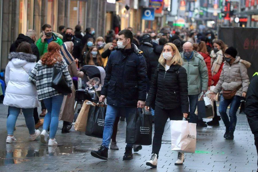Multitud de personas con mascarillas sanitarias caminan por una concurrida calle comercial cargando bolsas de tiendas.