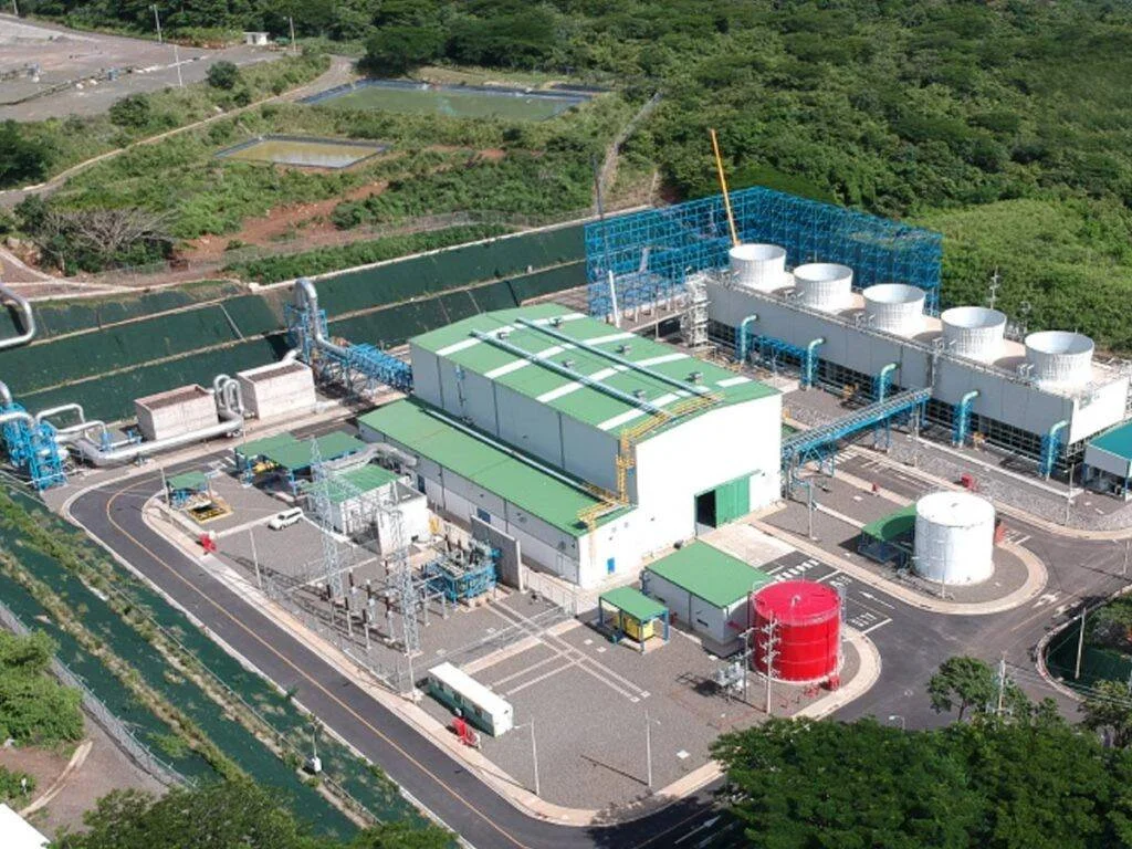 Vista aérea de una planta industrial de generación de energía con edificios de techo verde, torres de enfriamiento y tanques de almacenamiento, rodeada de vegetación.