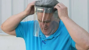 Un hombre con remera azul se coloca una pantalla facial protectora transparente sujetándola con ambas manos sobre su cabeza.