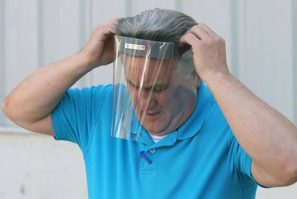 Un hombre con remera azul se coloca una pantalla facial protectora transparente sujetándola con ambas manos sobre su cabeza.